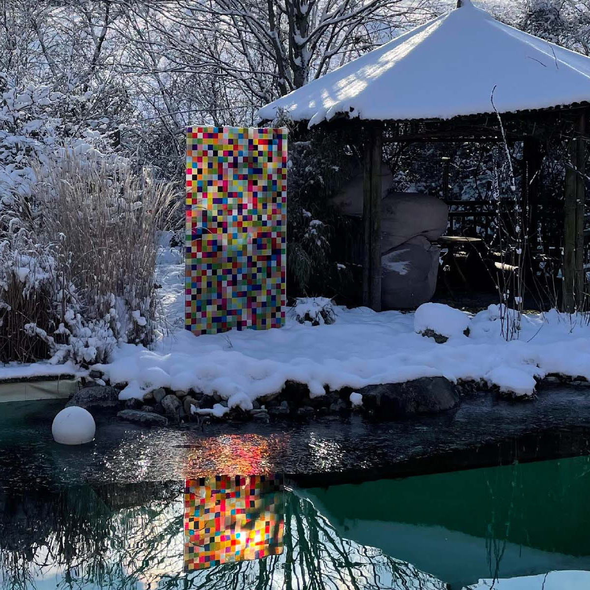 Die Glasdesign Stele "Pixel" der Künstlerin Pia Zeisel steht neben einer Hütte im Schnee und spiegelt sich in einem See.