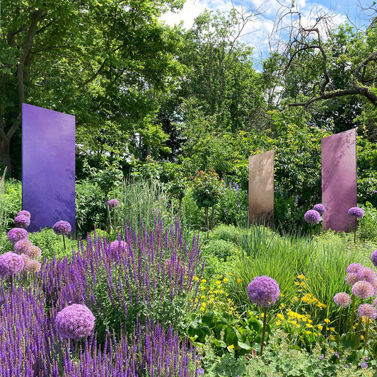 Ein violettes, ein blaues und ein beiges Glasdesign Element stehen in sehr üppiger Staudenbepflanzung mit perfekt harmonierenden Farbtönen. 