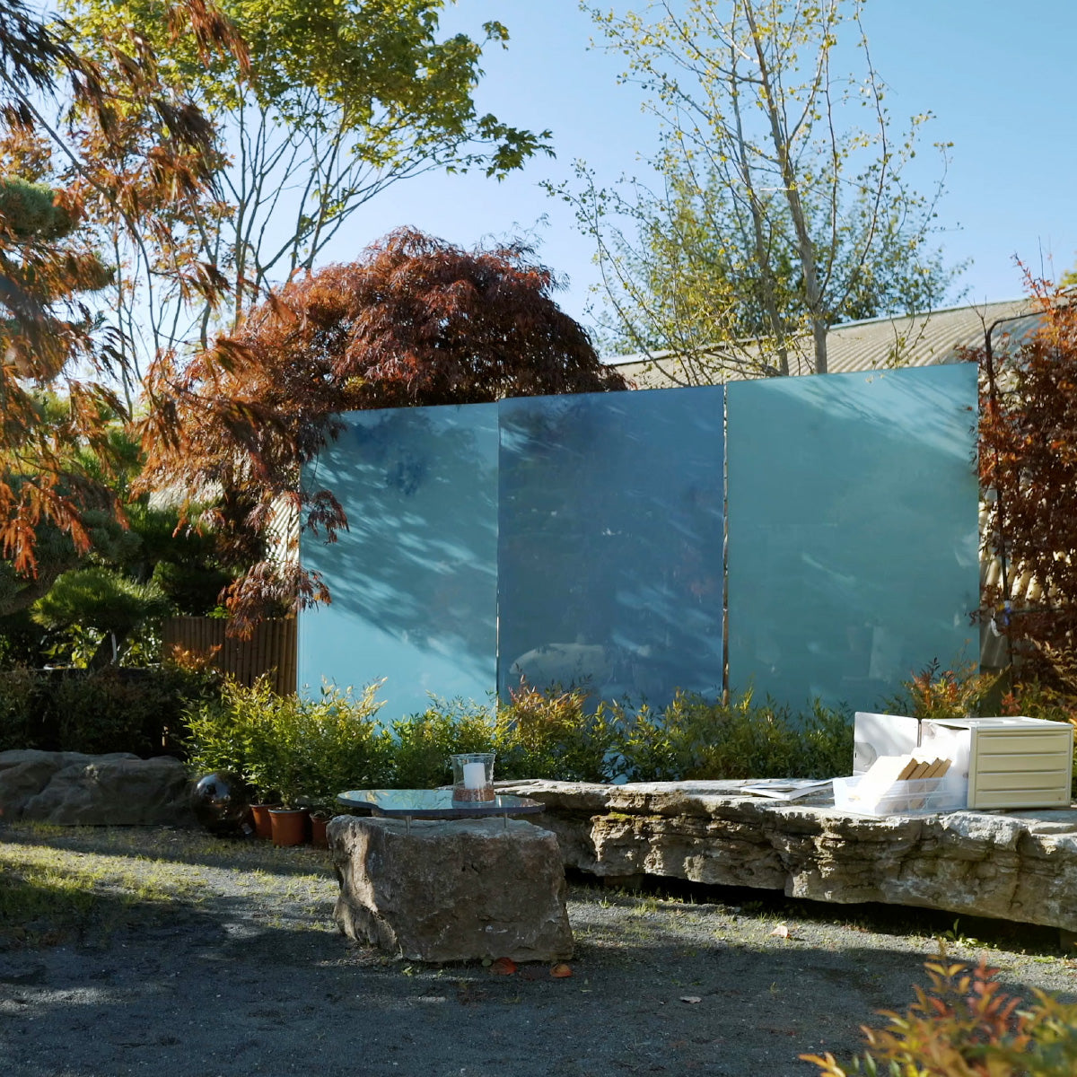 Drei blaue KNUMOX Glasdesign Elemente stehen in einem herbstlichen Garten mit Steinmauer.