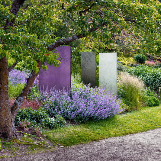 Eine violette, eine graue und eine weiße Glasdesign Stele stehen in der Pflanzung am Rand eines Weges in einem Park.