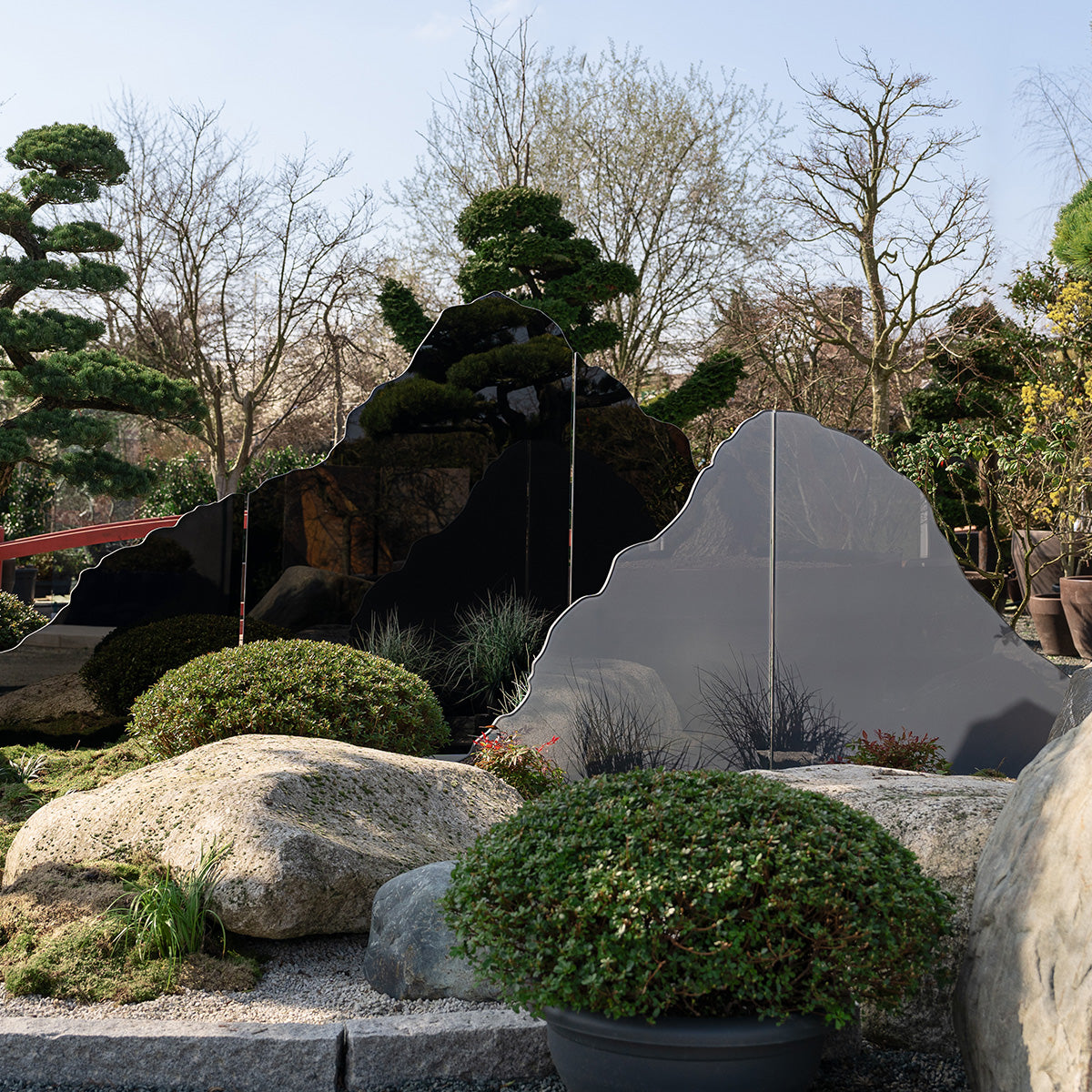 Die Serie "Mountain View" des Designers Marco Seidels steht in einem Garten mit Pflanzen vor Steinen. Die Glasdesign-Stelen sind in der Form einer Bergsilhouette ausgeschnitten. 