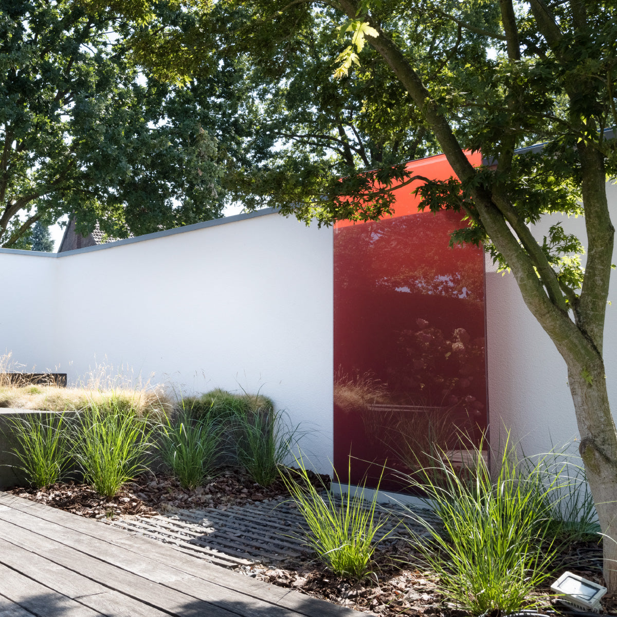 Das rote Design Element aus Glas steht vor einer Mauer in einem Garten, im Vordergrund ist ein Baum zu sehen. 