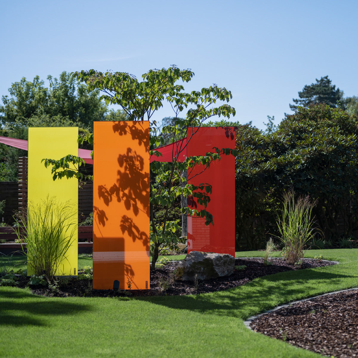 Ein gelbes, ein orangenes und ein rotes Sichtschutzelement stehen in einem Garten, man sieht die Schatten der Blätter auf dem mittleren Element. 