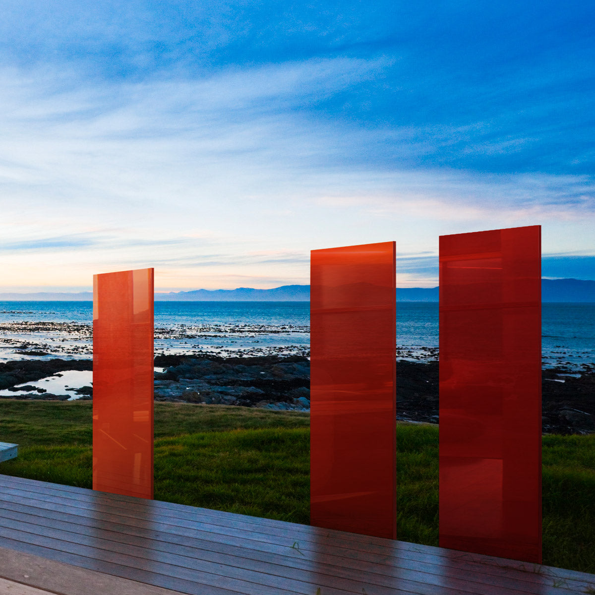 Drei Sichtschutz Glasdesignelemente in orange-roten farbabstufungen stehen an einer Terrasse mit Blick aufs Meer. 